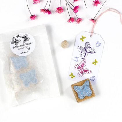 Stempel-Set Schmetterlinge von STUDIO KARAMELO: mehrere Holzstempel in transparenter Verpackung, daneben ein Geschenkanhänger mit bunten Stempelabdrücken in Pink, Lila und Grün